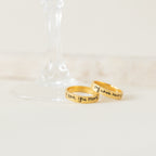 Two gold Handwriting Bands, engraved with "I love you more" and "my whole heart," rest by a wine glass base—ideal as a Christmas gift or personalized jewelry for someone special.