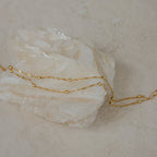 The Duo Bead Chain Bracelet pairs beautifully with a delicate gold chain necklace draped over a white crystal stone on a beige surface, creating an effortlessly chic and elegant look.