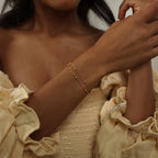 A woman in a cream ruffled dress wears the Duo Bead Chain Bracelet—two dainty bracelets featuring beaded and gold boho styles—on her wrist.