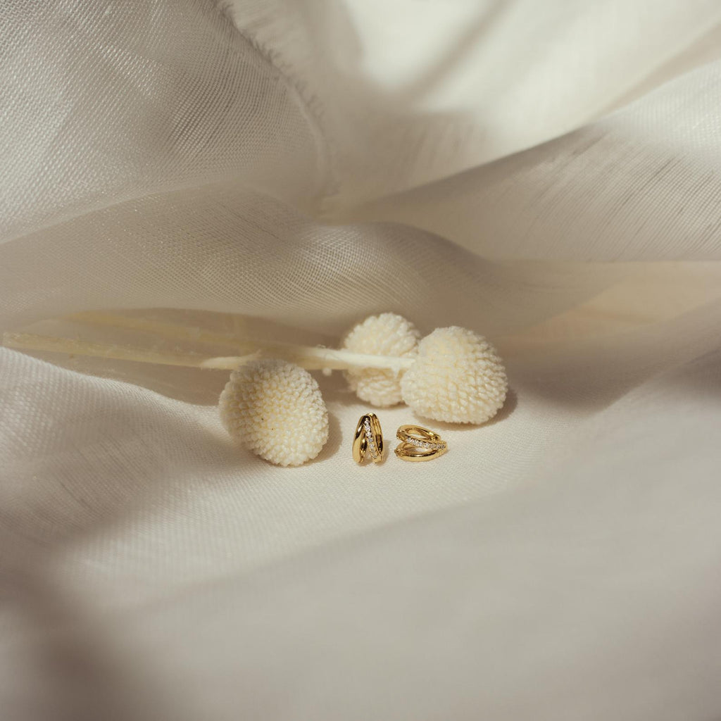 Pair of Amelia Duo Huggies resting near cream floral decor on satin fabric.