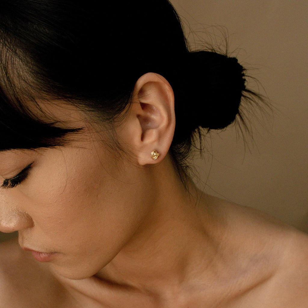 A woman with dark hair in a bun wears the Love Knot Studs, small gold earrings, as she looks downward with a neutral expression.