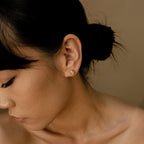 A woman with dark hair in a bun wears the Love Knot Studs, small gold earrings, as she looks downward with a neutral expression.