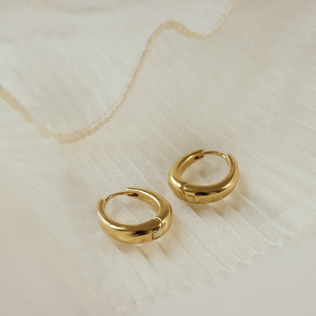 The Paris Oval Hoops, classic gold earrings, are displayed elegantly on a white, pleated fabric background.