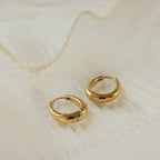 The Paris Oval Hoops, classic gold earrings, are displayed elegantly on a white, pleated fabric background.