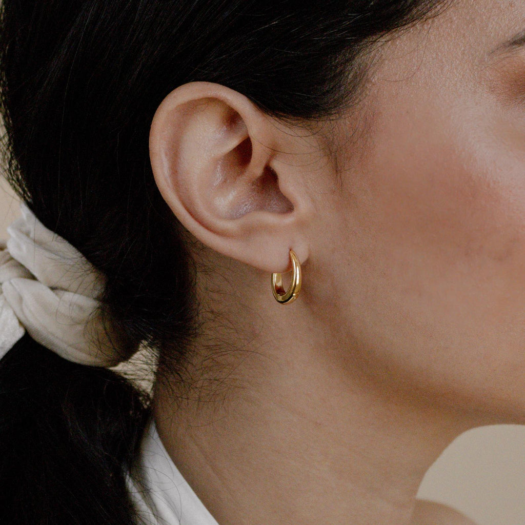 Close-up of a woman's ear wearing Paris Oval Hoops in 18K Gold, her dark hair tied back with a light-colored scarf.