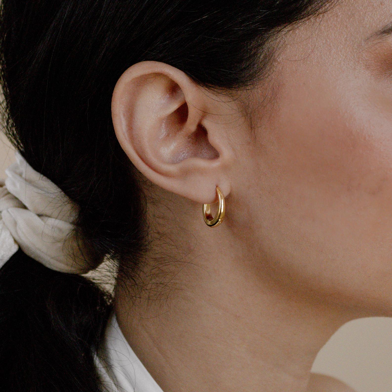 Close-up of a woman's ear wearing Paris Oval Hoops in 18K Gold, her dark hair tied back with a light-colored scarf.