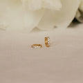 The Elise Diamond Huggies, small pave earrings adorned with sparkling stones, are displayed on soft beige fabric with white flowers in the background.