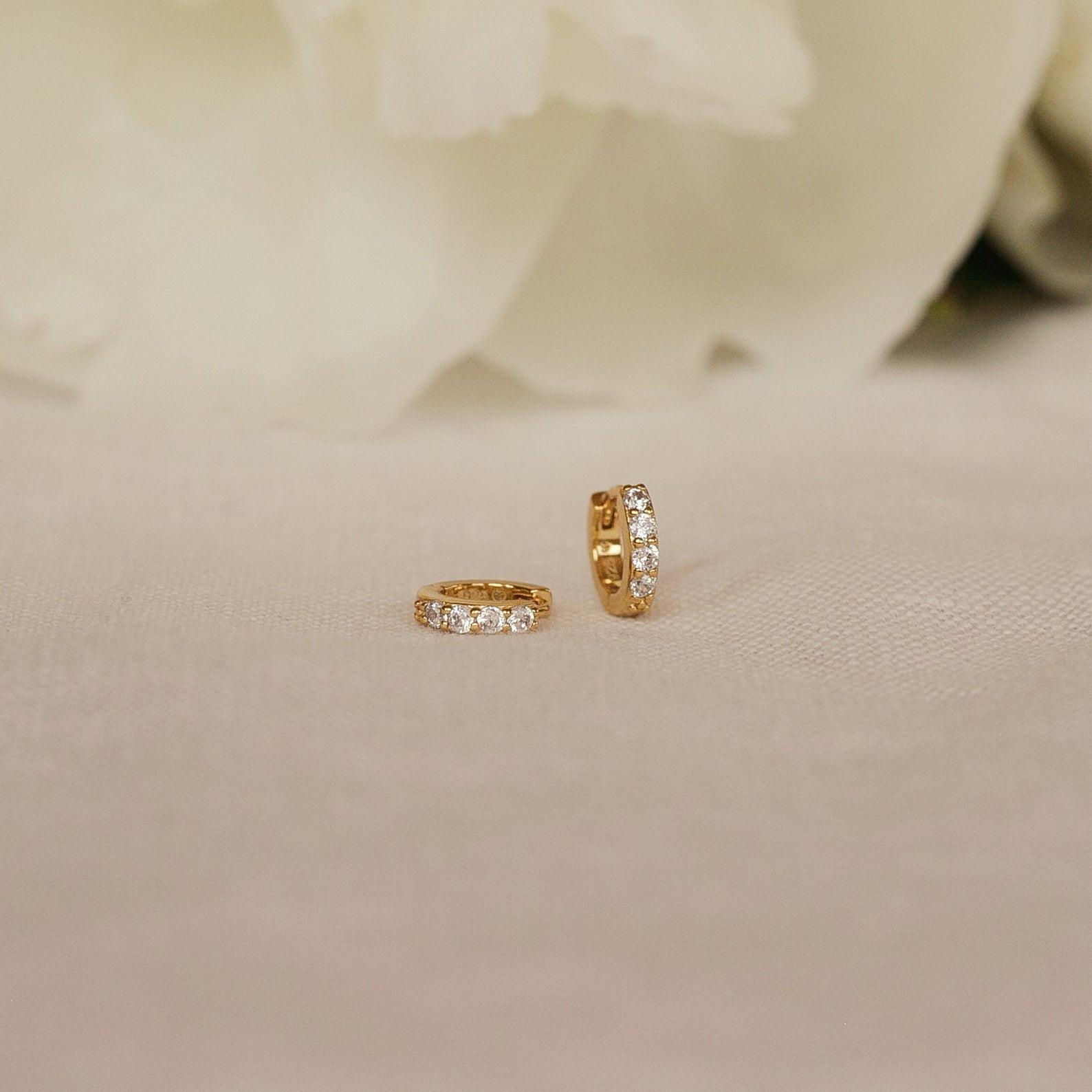 The Elise Diamond Huggies, small pave earrings adorned with sparkling stones, are displayed on soft beige fabric with white flowers in the background.