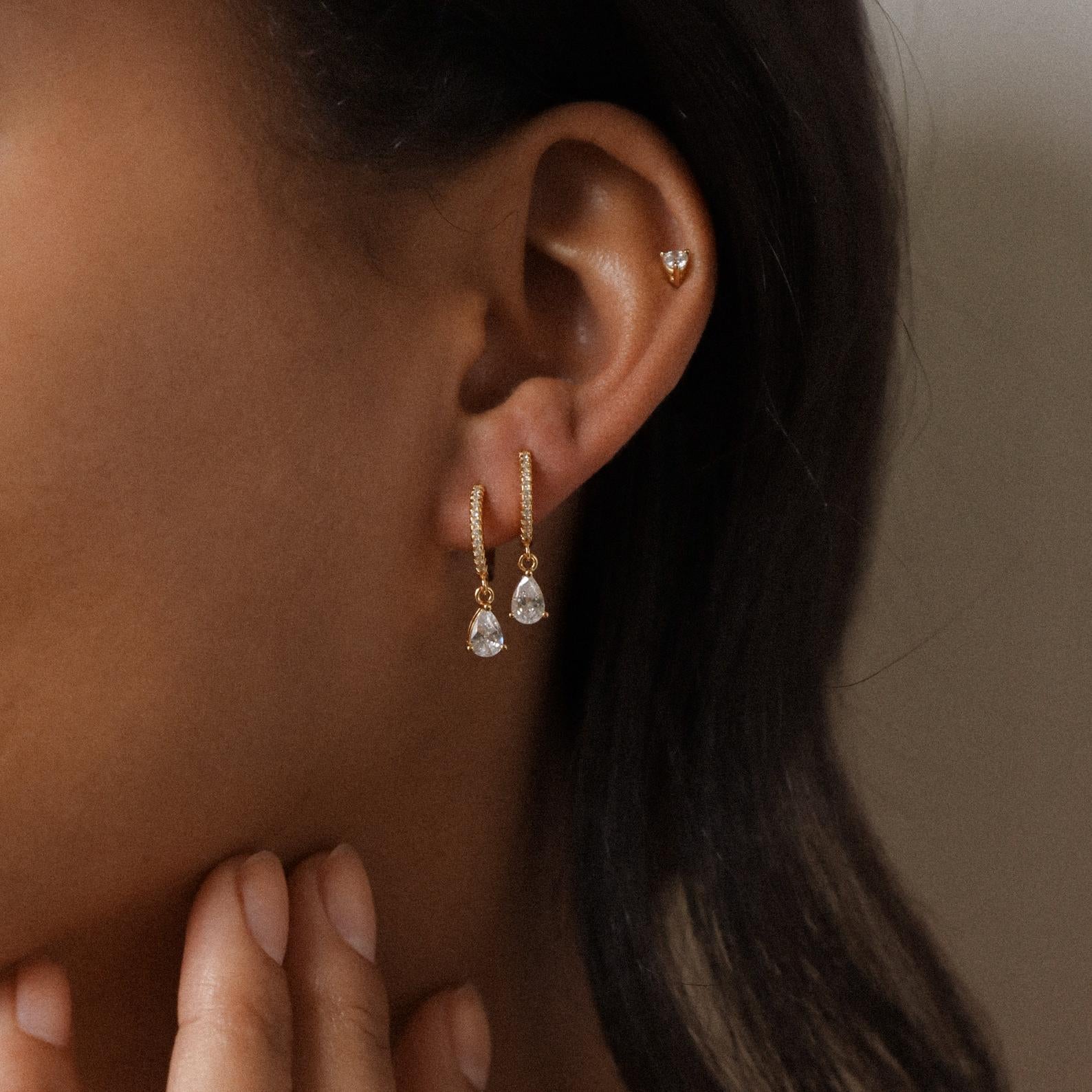 A woman wears Diamond Drop Huggies—gold hoop earrings with diamond accents and clear teardrop pendants—plus a small heart stud on her upper ear.