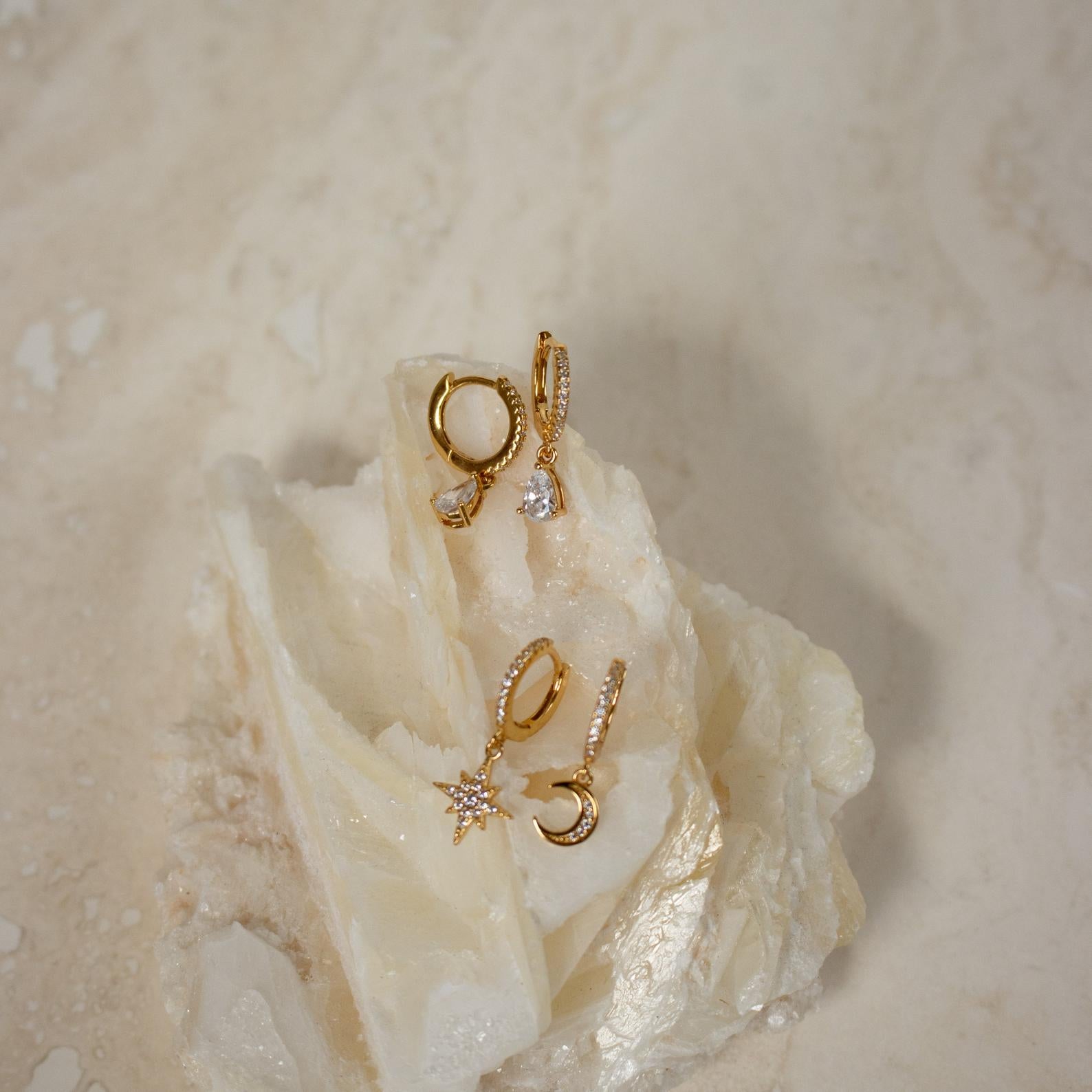 Three pairs of gold earrings, featuring Diamond Drop Huggies and semi-formal styles with crystal charms, are displayed on a white textured stone.
