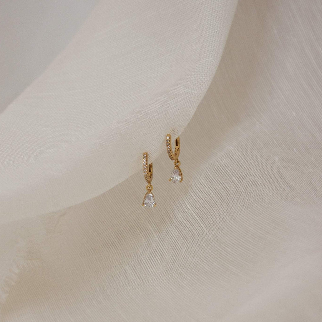 A pair of elegant gold drop earrings with pave crystals along the hoops and teardrop-shaped clear gemstones hanging from each. The earrings are delicately displayed against a soft, cream-colored fabric backdrop. 