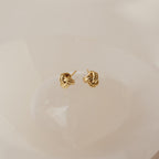 Love Knot Studs in a small gold design displayed on a smooth, light-colored surface—perfect as symbolic jewelry or a thoughtful gift.
