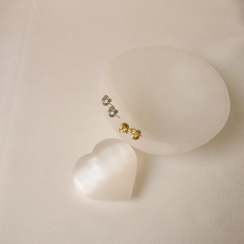 The Love Knot Studs in gold and silver sit on an elegant oval stone, with a shimmering white heart nearby—symbolic jewelry that makes a thoughtful gift for someone special.