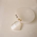 The Love Knot Studs in gold and silver sit on an elegant oval stone, with a shimmering white heart nearby—symbolic jewelry that makes a thoughtful gift for someone special.