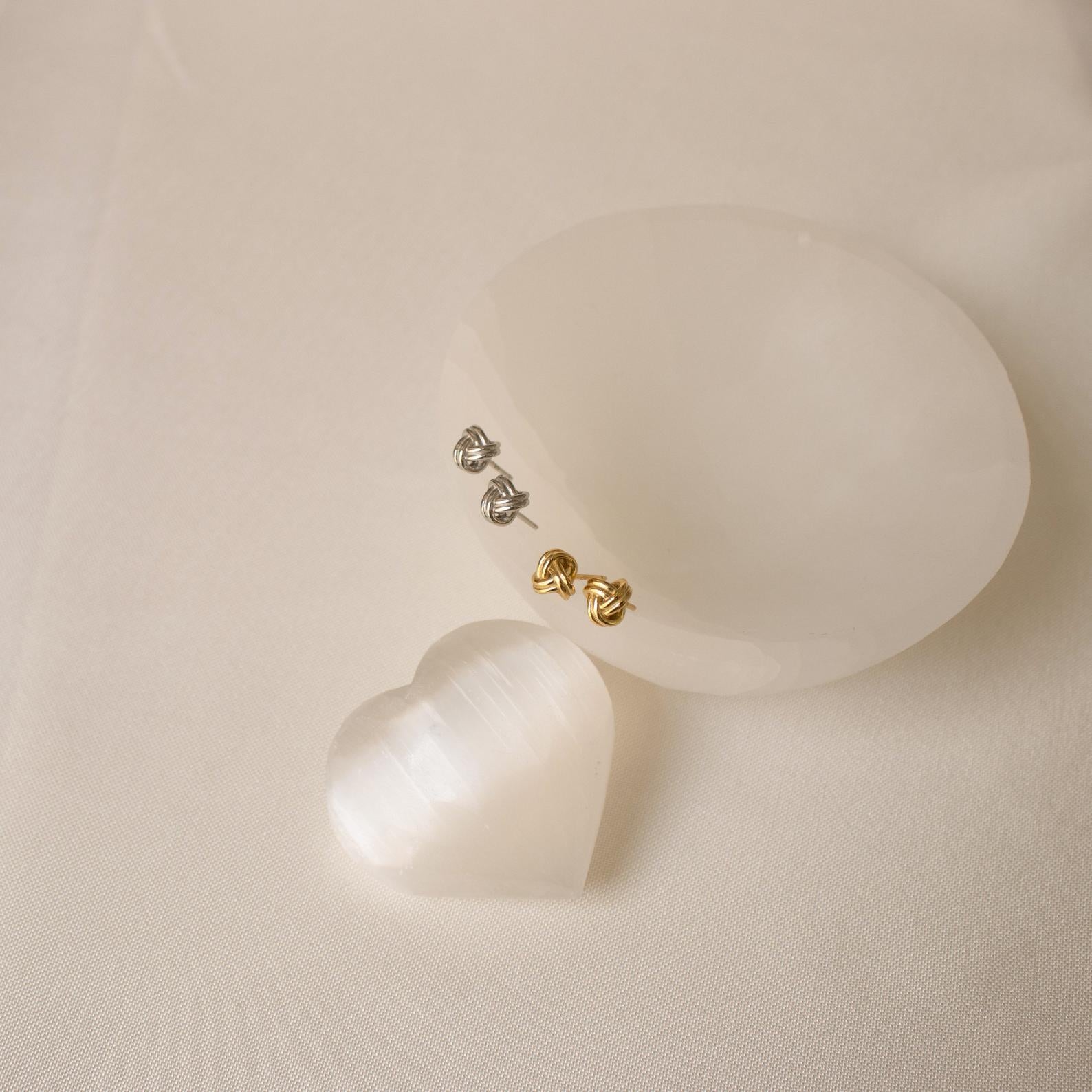 The Love Knot Studs in gold and silver sit on an elegant oval stone, with a shimmering white heart nearby—symbolic jewelry that makes a thoughtful gift for someone special.