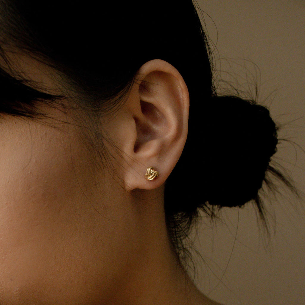 Close-up of a woman's ear wearing Love Knot Studs, small gold earrings symbolizing love—perfect as a thoughtful gift. Her dark hair is tied in a low bun.