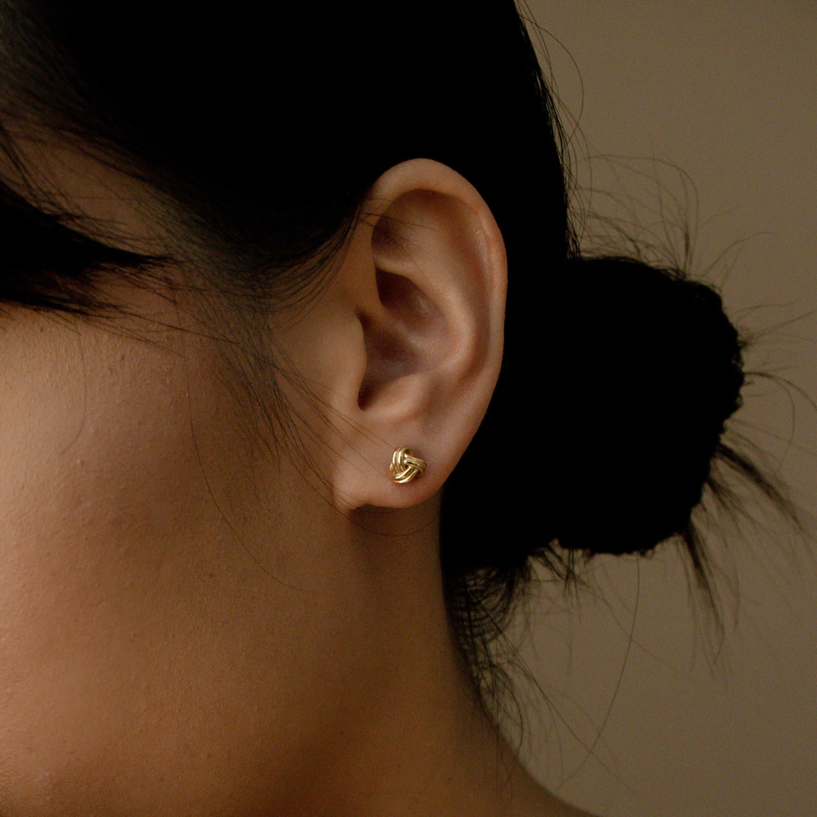 Close-up of a woman's ear wearing Love Knot Studs, small gold earrings symbolizing love—perfect as a thoughtful gift. Her dark hair is tied in a low bun.