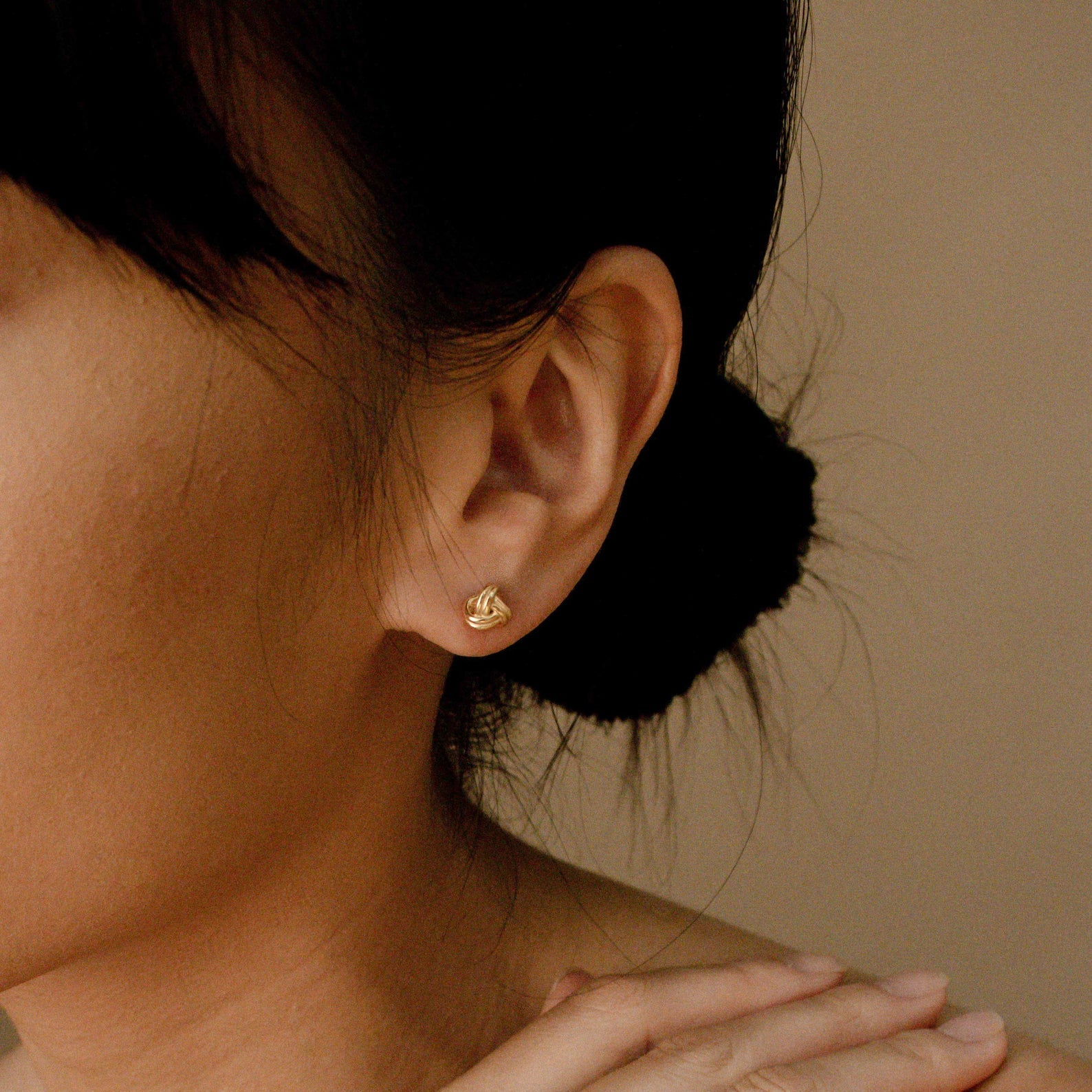 A woman with dark hair in a bun wears Love Knot Studs, elegant and symbolic jewelry, her hand gently placed near her neck.