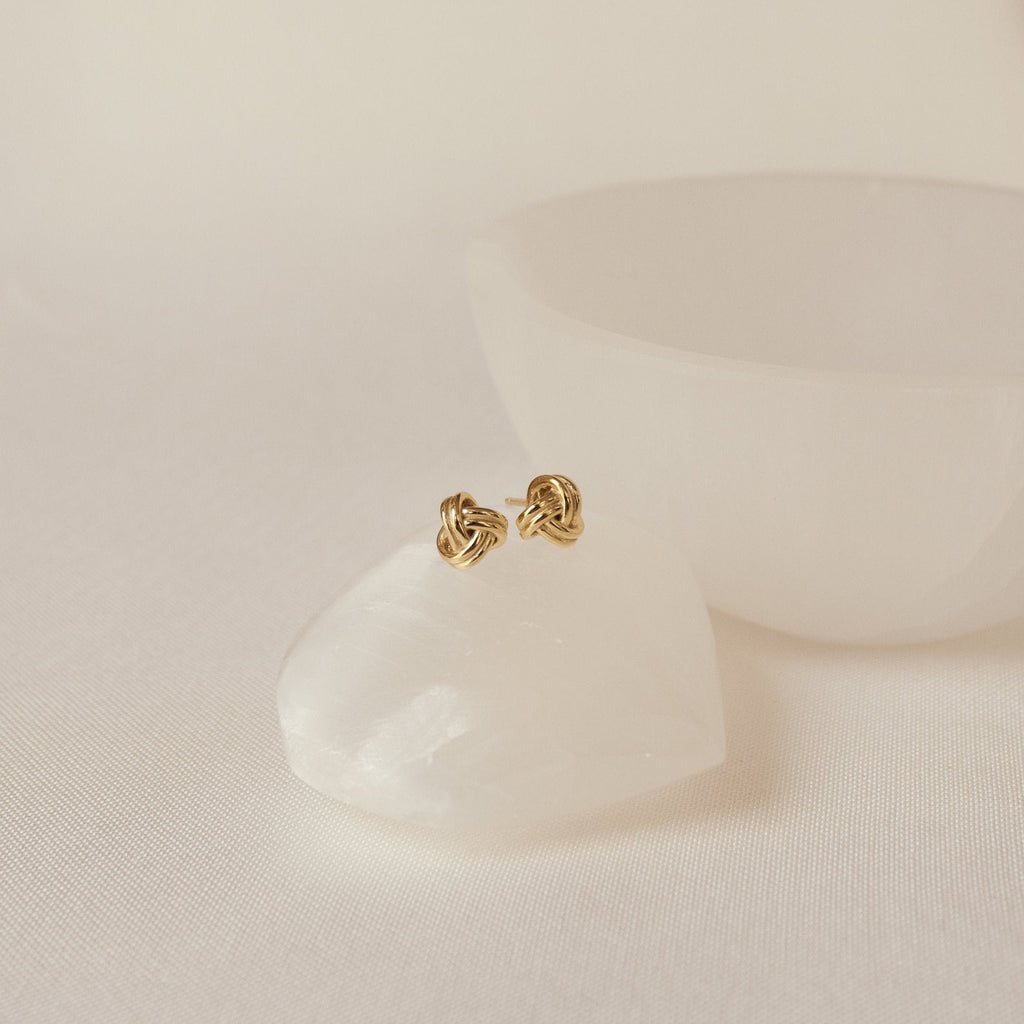 Love Knot Studs rest on a smooth white stone with a white bowl behind them. These earrings symbolize timeless connection and elegance, making them a thoughtful gift.