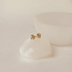 Love Knot Studs rest on a smooth white stone with a white bowl behind them. These earrings symbolize timeless connection and elegance, making them a thoughtful gift.