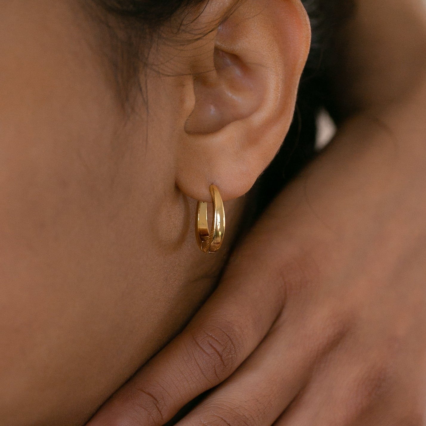 Close-up of the Paris Oval Hoops gold earring on an ear, accented by a hand gently resting on the neck—an elegant, classic-inspired accessory.
