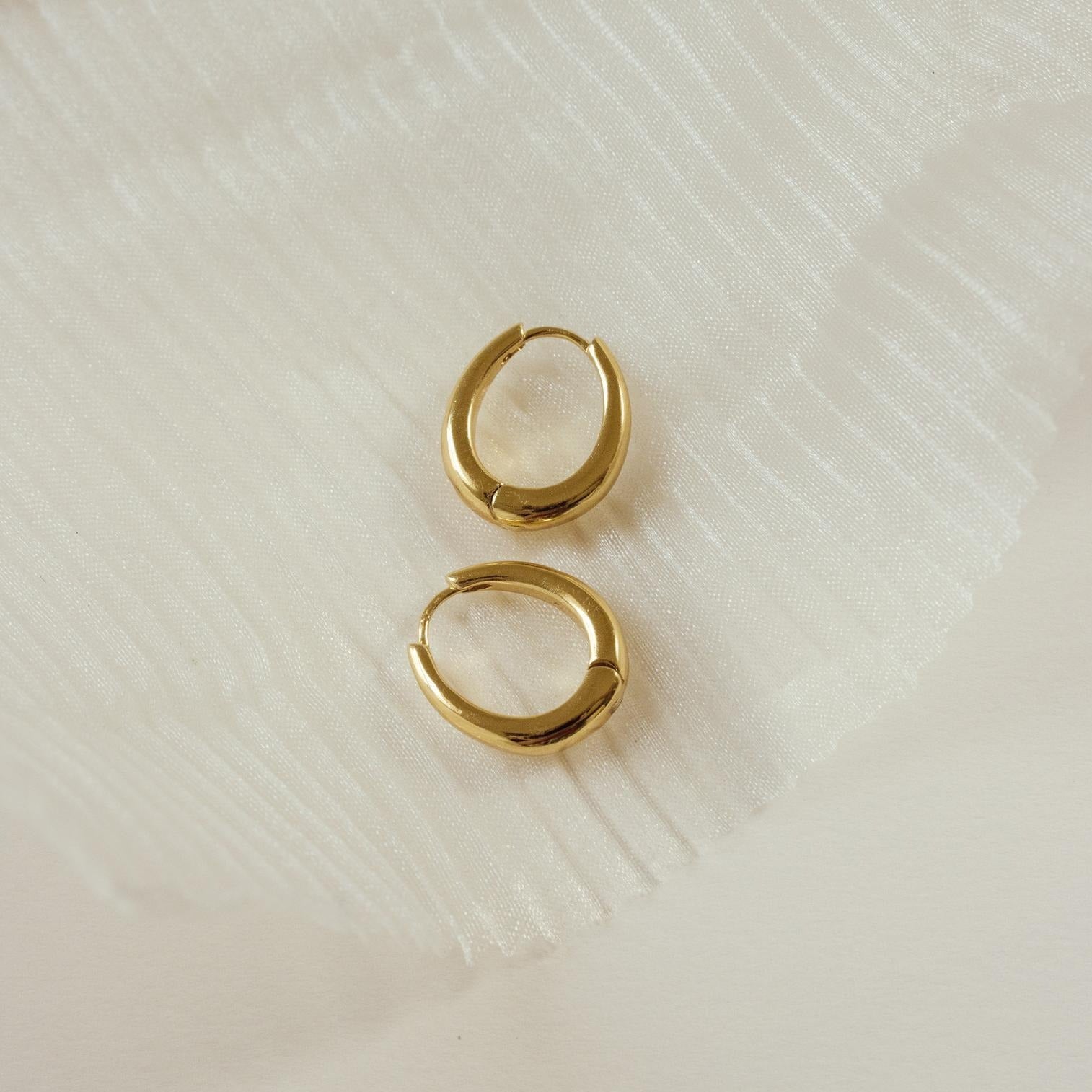 The Paris Oval Hoops earrings are displayed on a textured white fabric background.