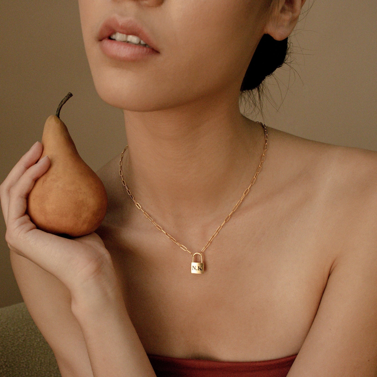 A woman holding a pear wears the Padlock Paperclip Necklace—a gold padlock charm engraved "N A" on a stylish paperclip chain.