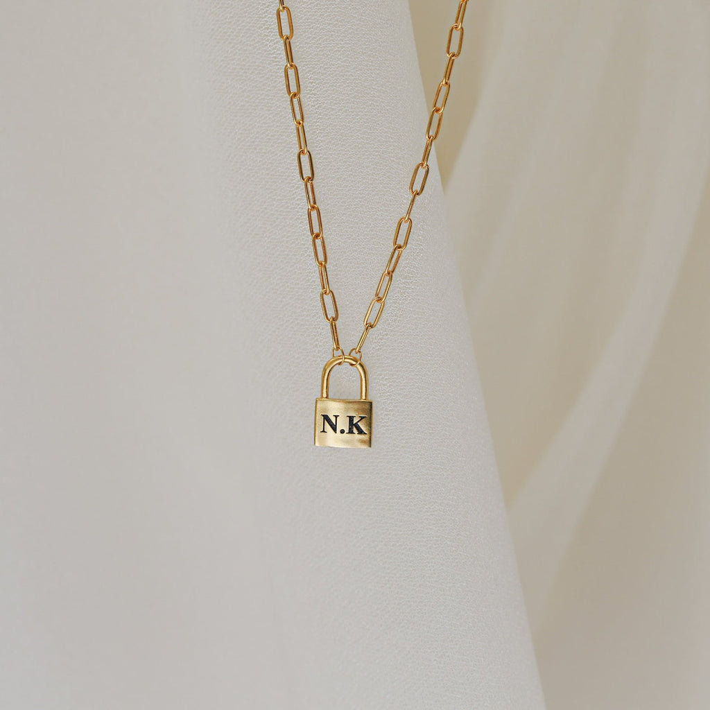 The Padlock Paperclip Necklace, engraved with the initials N.K., shines on a soft cream background. This elegant gold chain charm necklace brings a modern touch to any style.