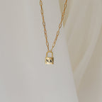 The Padlock Paperclip Necklace, engraved with the initials N.K., shines on a soft cream background. This elegant gold chain charm necklace brings a modern touch to any style.