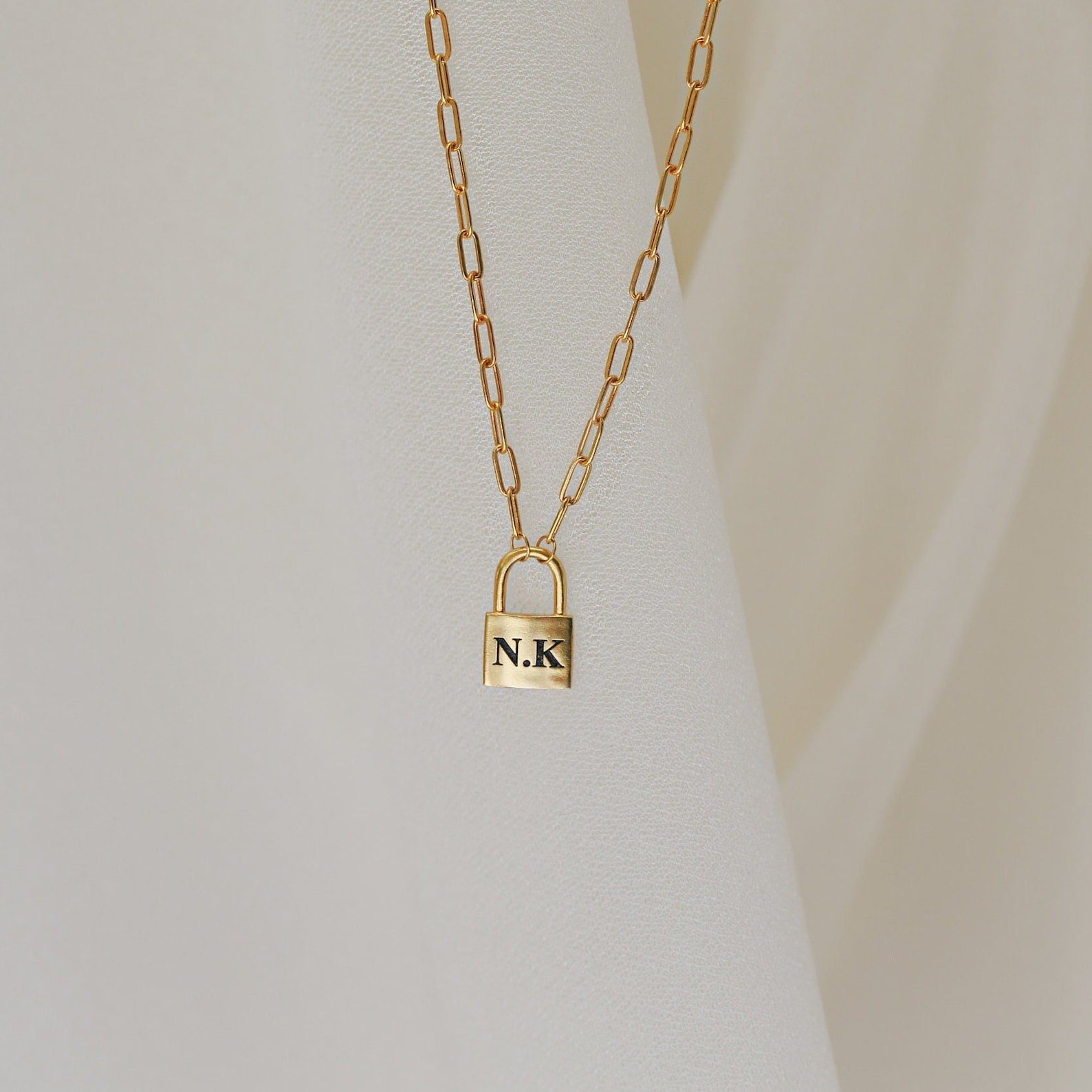 The Padlock Paperclip Necklace, engraved with the initials N.K., shines on a soft cream background. This elegant gold chain charm necklace brings a modern touch to any style.