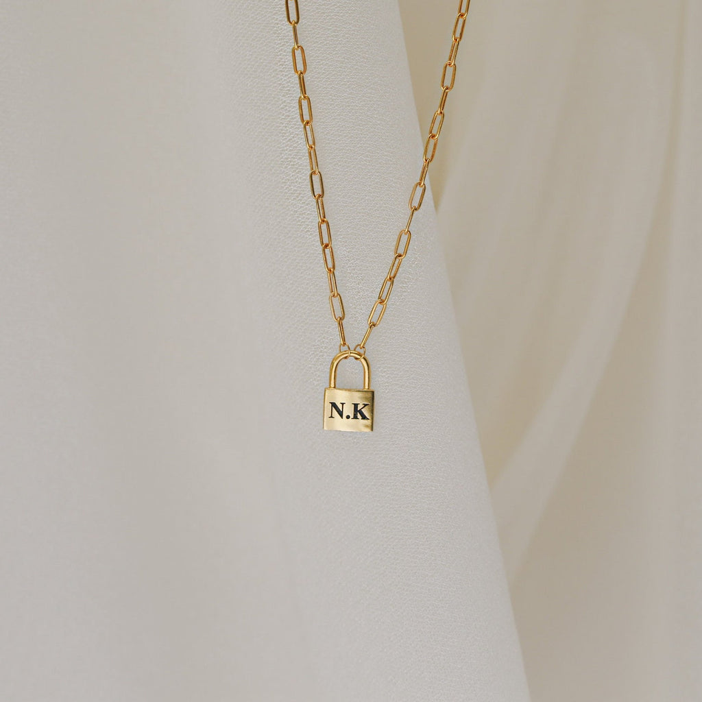 The Padlock Paperclip Necklace, featuring a gold padlock pendant engraved with “N.K.,” rests elegantly against a cream fabric background.