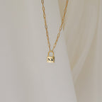 The Padlock Paperclip Necklace, featuring a gold padlock pendant engraved with “N.K.,” rests elegantly against a cream fabric background.