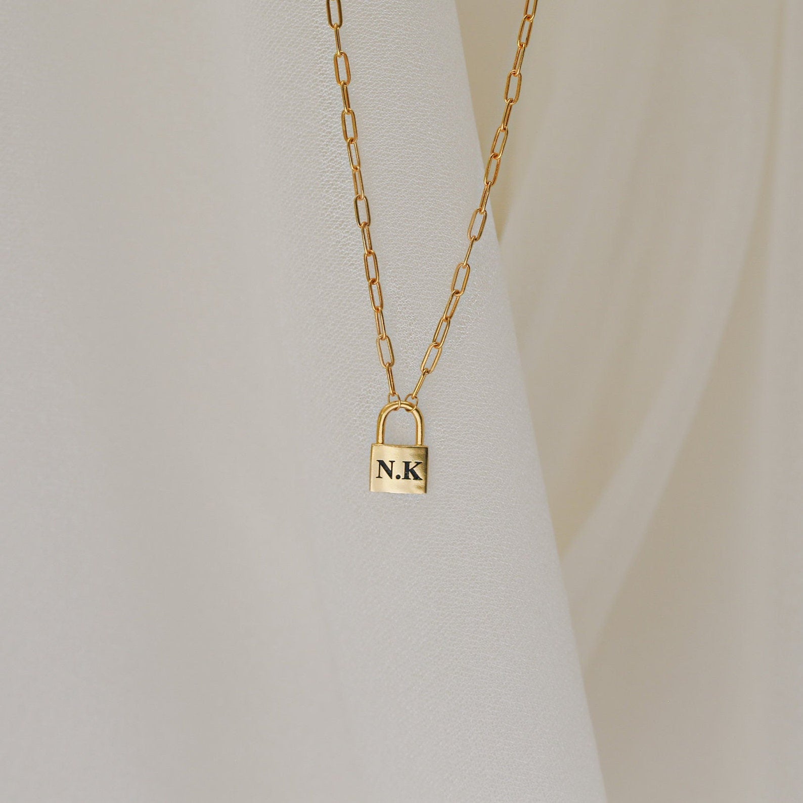 The Padlock Paperclip Necklace, featuring a gold padlock pendant engraved with “N.K.,” rests elegantly against a cream fabric background.