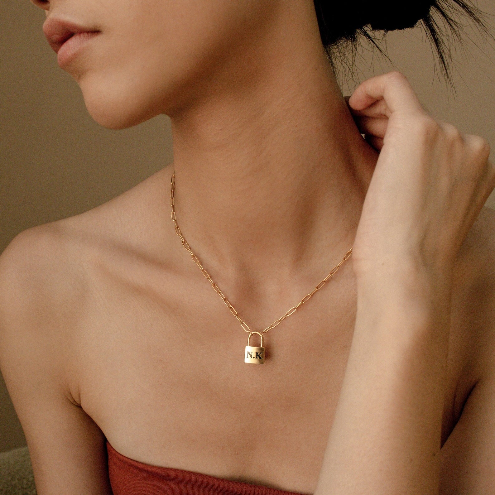 A woman touches her neck while wearing the Padlock Paperclip Necklace, featuring a gold pendant engraved with N.K.