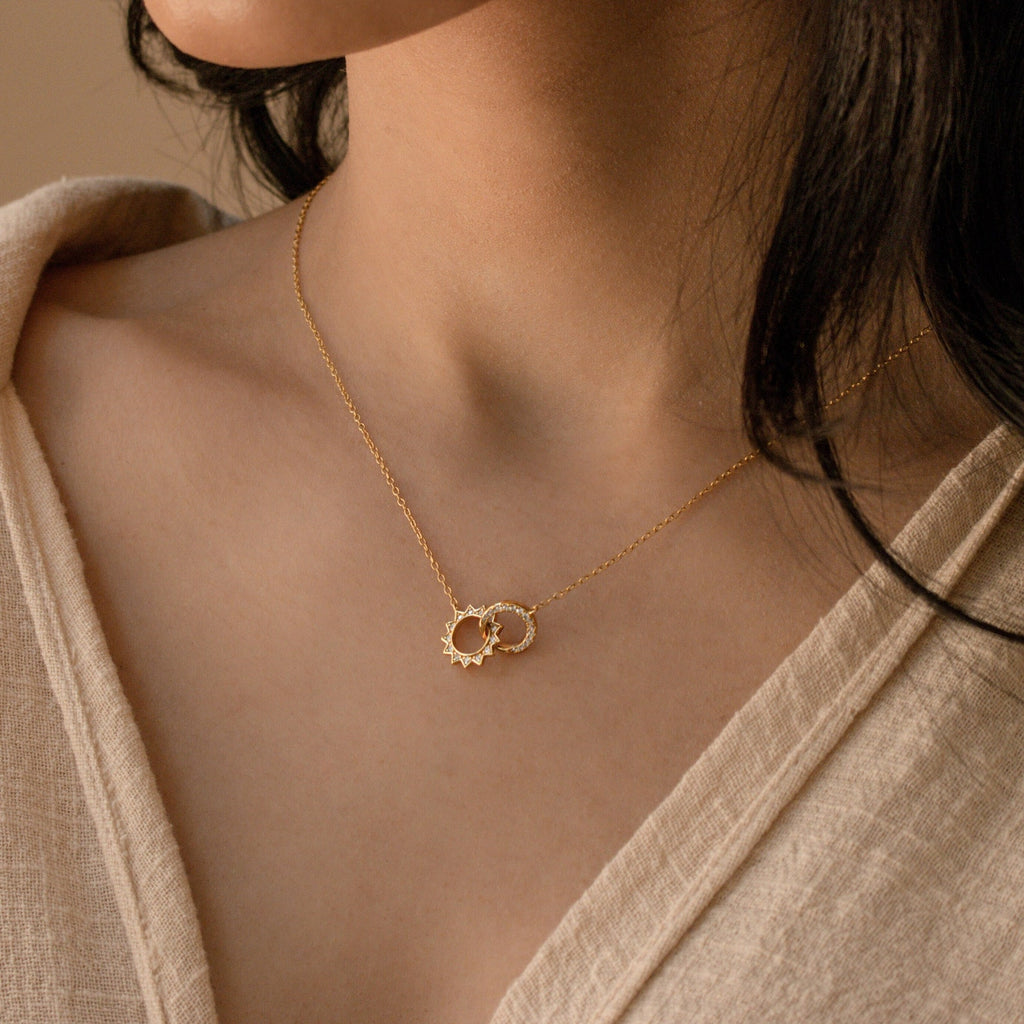 A woman wears the Sun  Moon Eternity Necklace, featuring two interlocking celestial pendants, elegantly paired with a beige top.