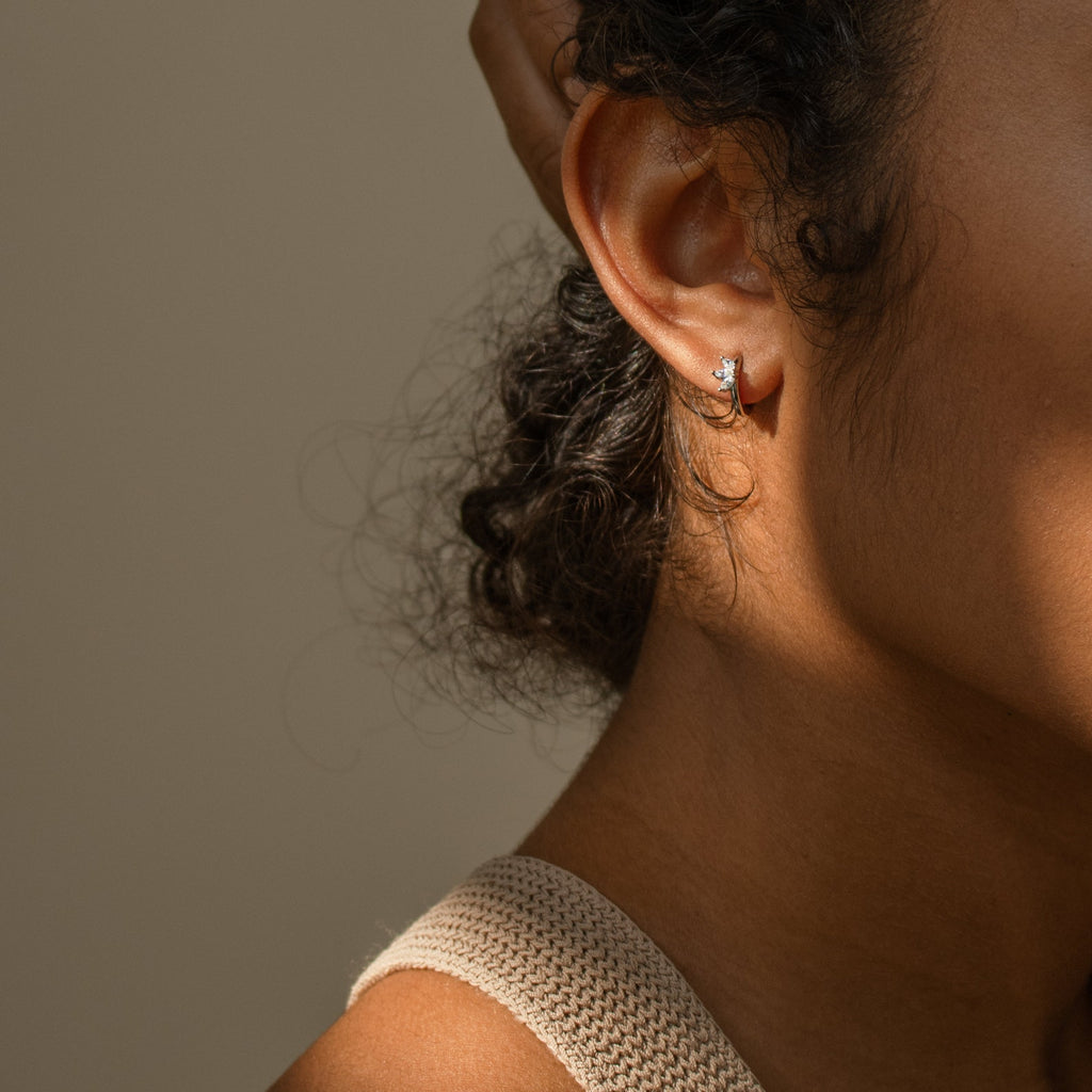 Close-up of a woman's ear wearing Giana Marquise Huggies—a subtle, elegant earring—her hand gently resting on her head in soft lighting.