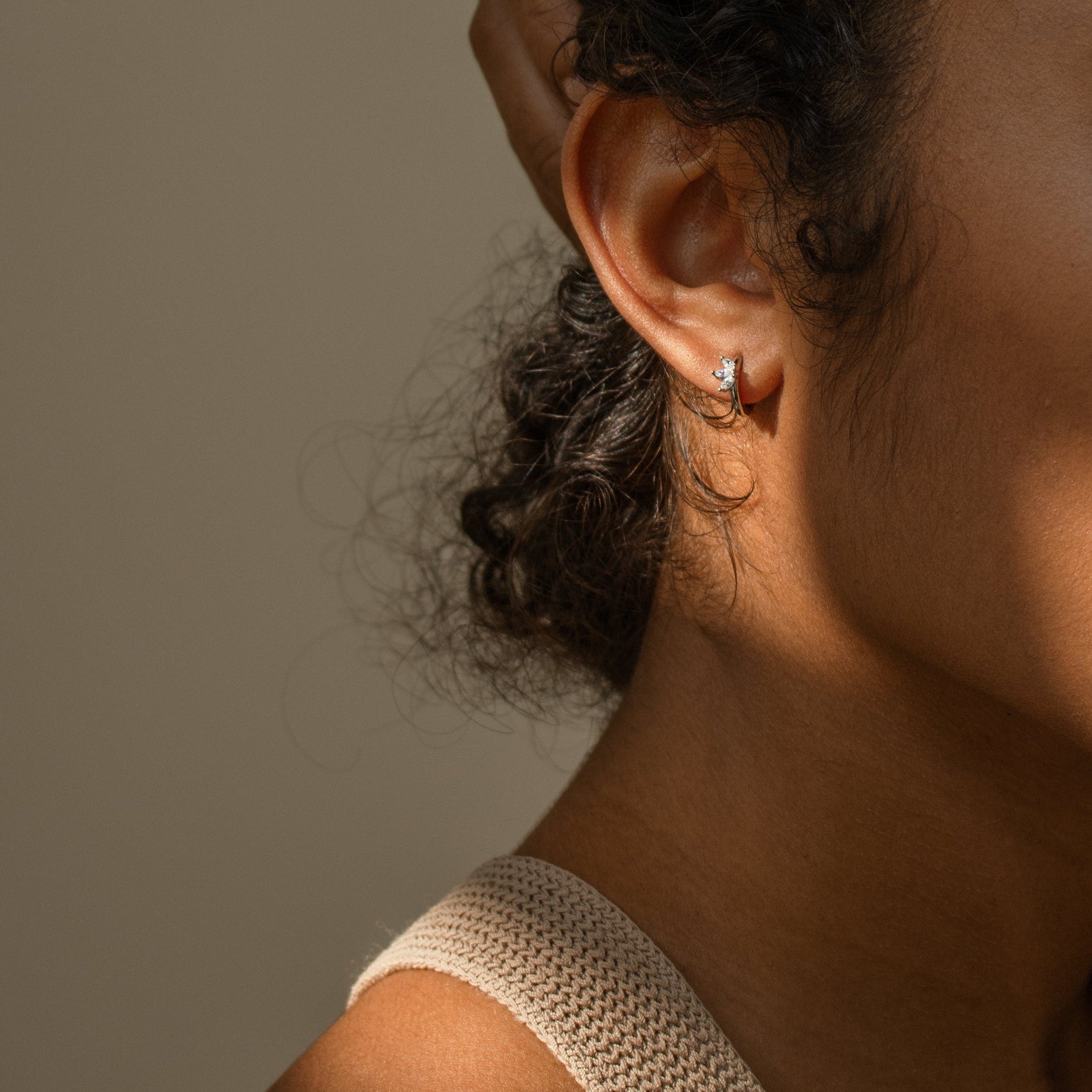 Close-up of a woman's ear wearing Giana Marquise Huggies—a subtle, elegant earring—her hand gently resting on her head in soft lighting.