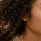 Close-up of a woman’s ear with curly hair, wearing the Giana Marquise Huggies—delicate gold hoops with a chic charm, ideal for lovers of understated style.