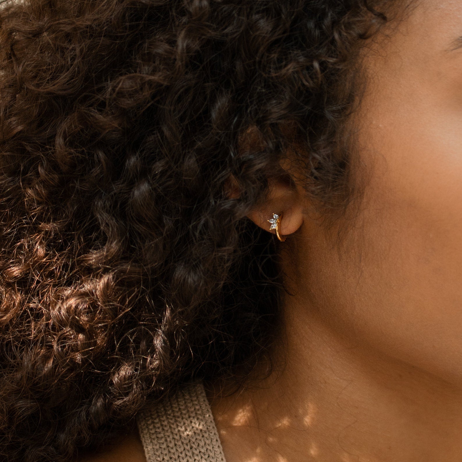 Close-up of a woman’s ear with curly hair, wearing the Giana Marquise Huggies—delicate gold hoops with a chic charm, ideal for lovers of understated style.