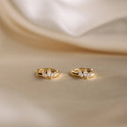 Two gold rings with three marquise-cut diamonds each, displayed on a soft beige fabric background, create an elegant look when paired with subtle earrings like the Giana Marquise Huggies.