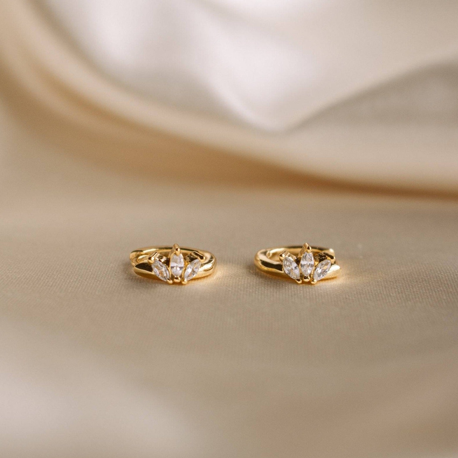 Two gold rings with three marquise-cut diamonds each, displayed on a soft beige fabric background, create an elegant look when paired with subtle earrings like the Giana Marquise Huggies.