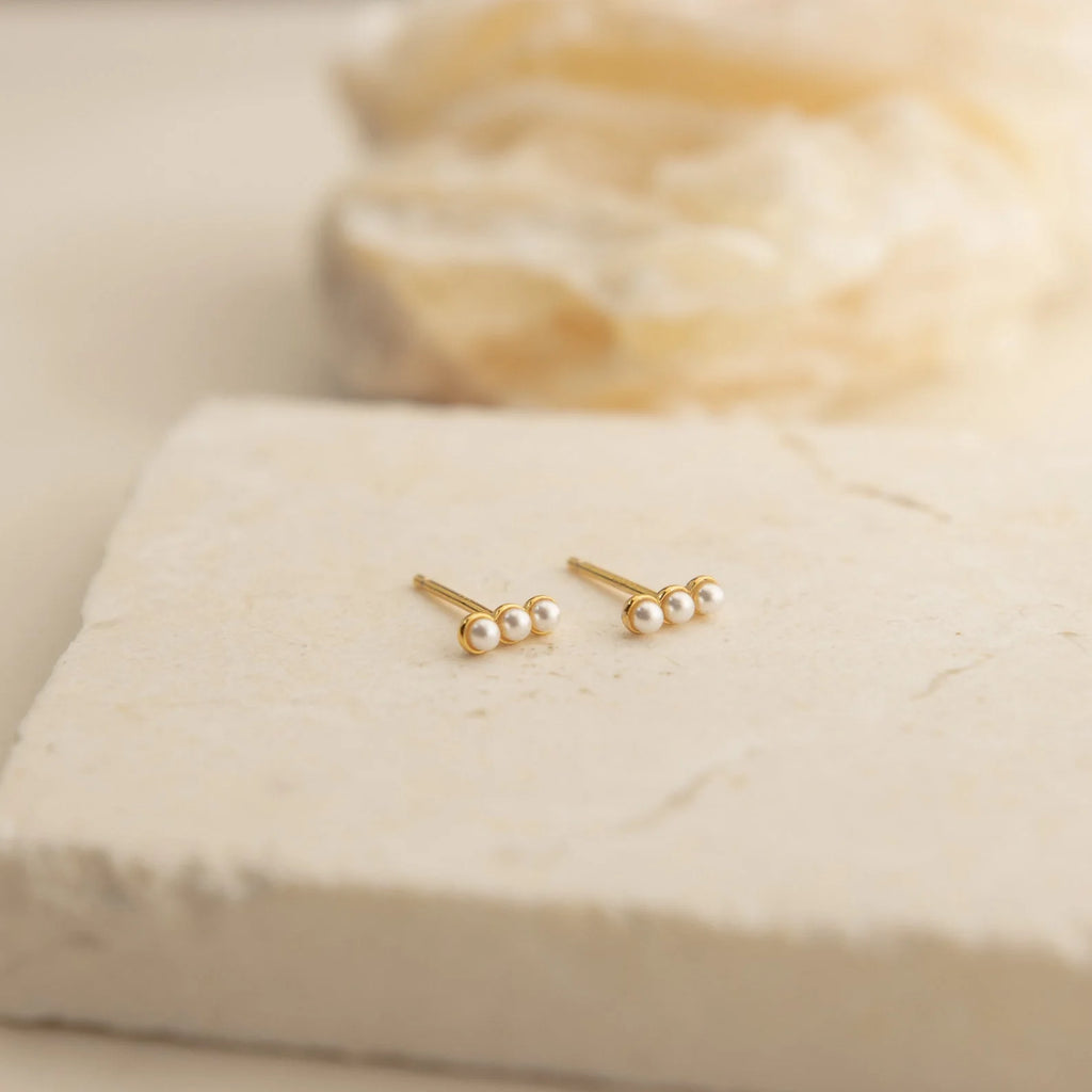 The Pearl Earrings Set features gold studs with three small pearls on each, beautifully displayed on a textured light stone surface—a timeless addition to your collection.