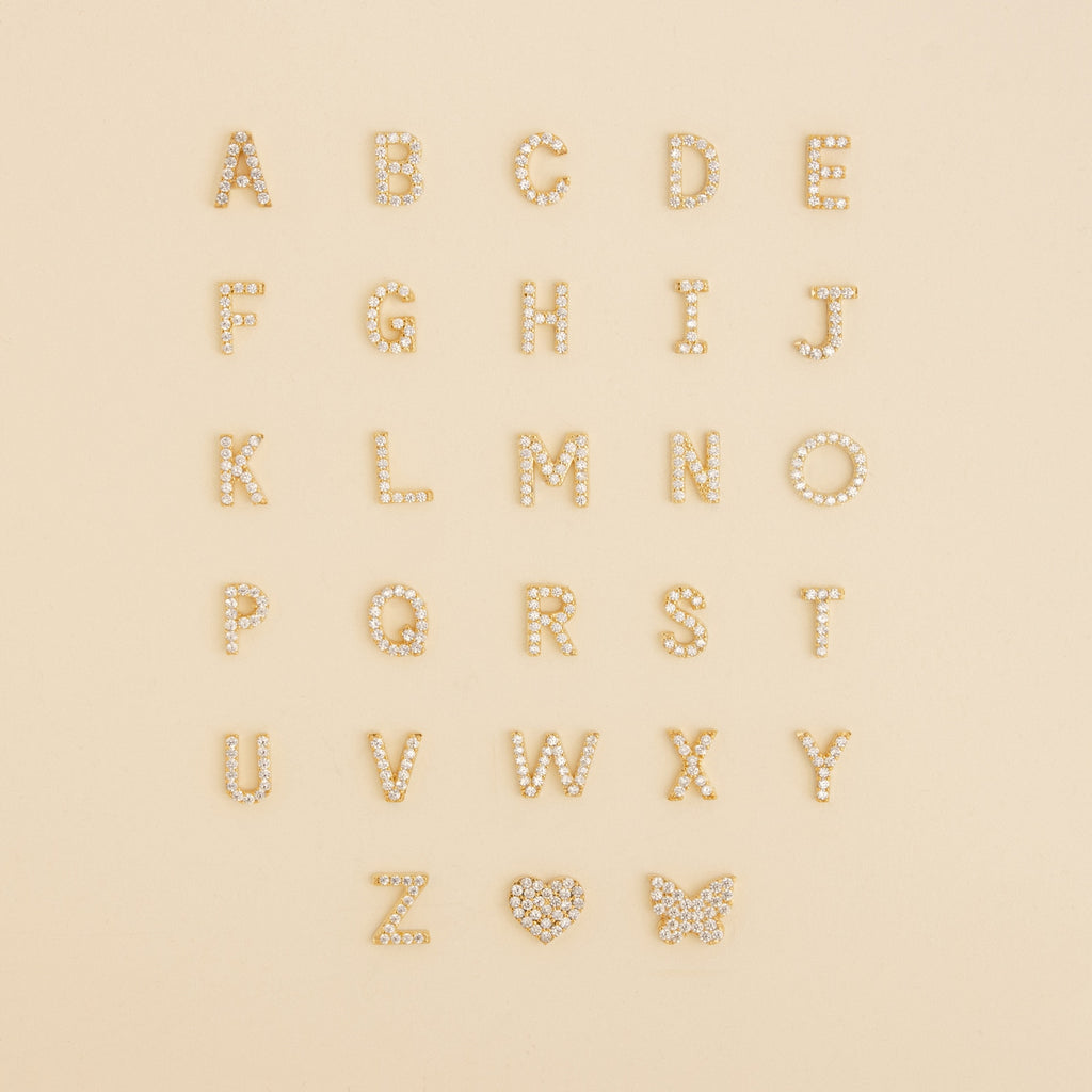 Pave Letter Studs A-Z arranged in rows with a sparkling rhinestone heart and butterfly on a beige background—ideal for creating personalized earrings or adding a touch of minimal style.
