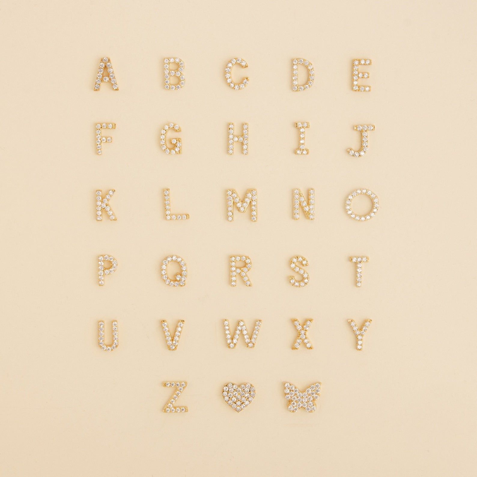 Pave Letter Studs A-Z arranged in rows with a sparkling rhinestone heart and butterfly on a beige background—ideal for creating personalized earrings or adding a touch of minimal style.
