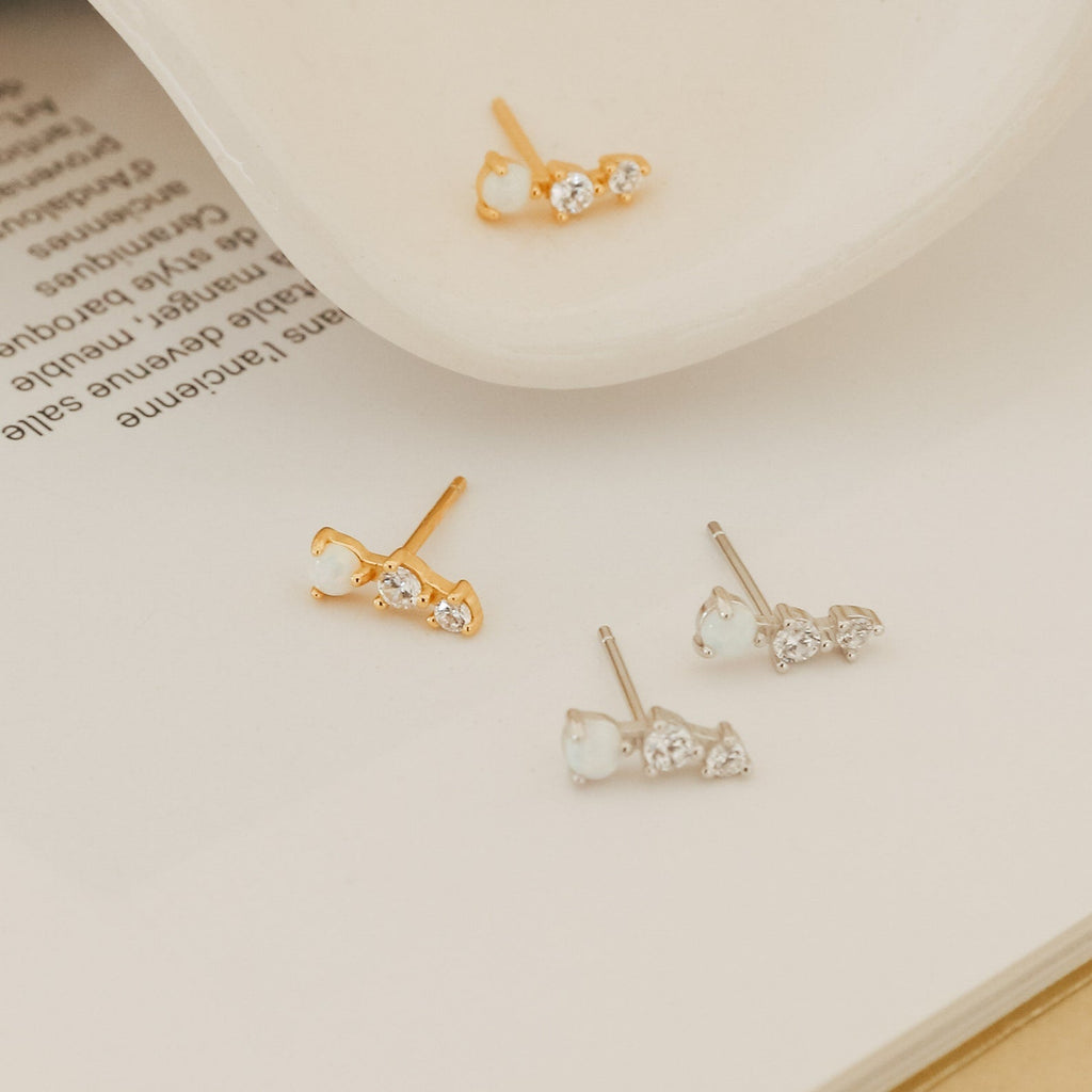 The Dainty Opal Earrings Set features two pairs of delicate earrings—one gold, one silver—each adorned with three small opals, beautifully displayed on a book for everyday elegance.