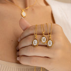 A hand holds three gold enamel Initial Pendant Necklaces—each with an L, M, or R pendant; another vintage-inspired necklace is worn around the neck.
