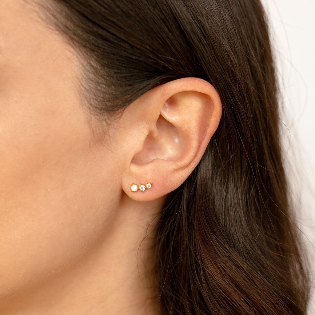 Close-up of a woman's ear adorned with three delicate earrings, featuring the Dainty Opal Earrings Set with differently colored stones.