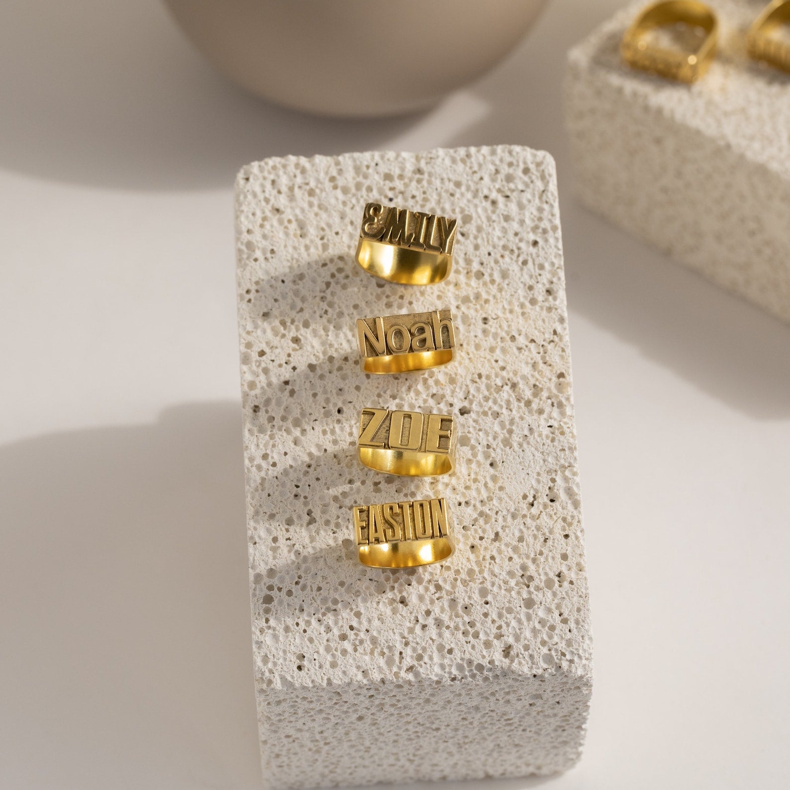 Four Bold Name Rings engraved with Emily, Noah, Zoe, and Easion displayed on a textured stone block—a striking personalized jewelry collection perfect for making a statement.