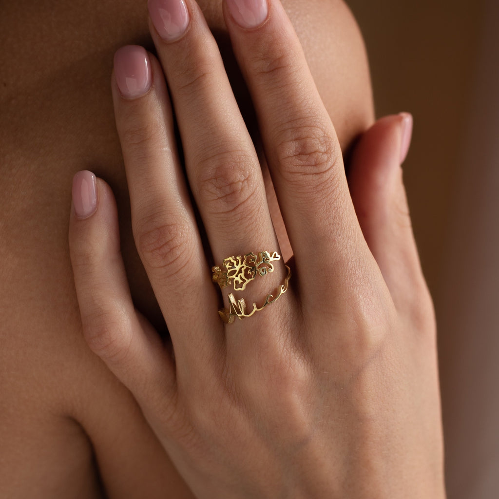 A hand with light pink nails wears a gold Flower Name Ring featuring a tree design and script—an elegant gift for moms or a unique custom flower jewelry piece.