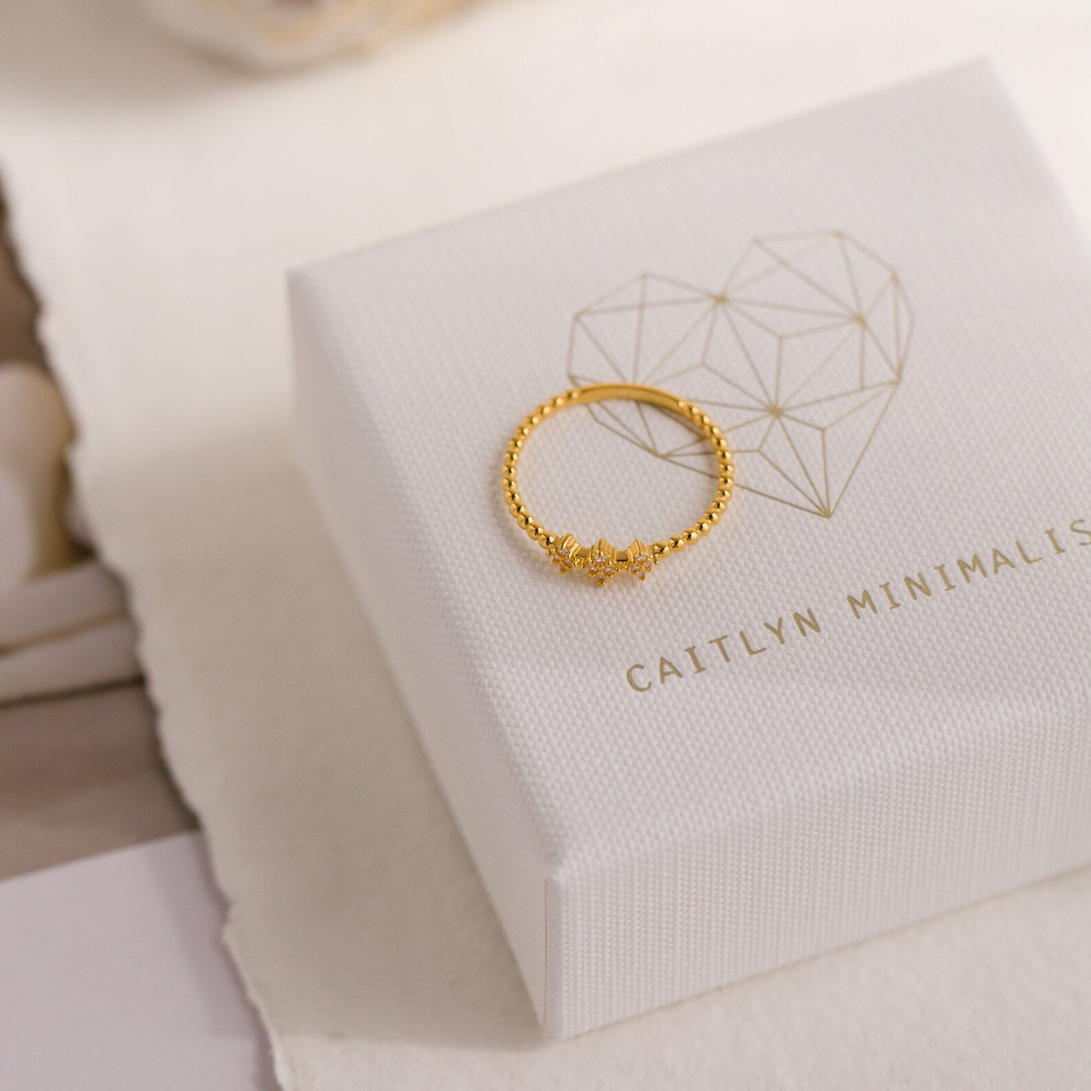 The Jasmine Beaded Diamond Ring sits atop a white jewelry box featuring a geometric heart and Caitlyn Minimalist text, making it a perfect addition to your favorite ring stack.